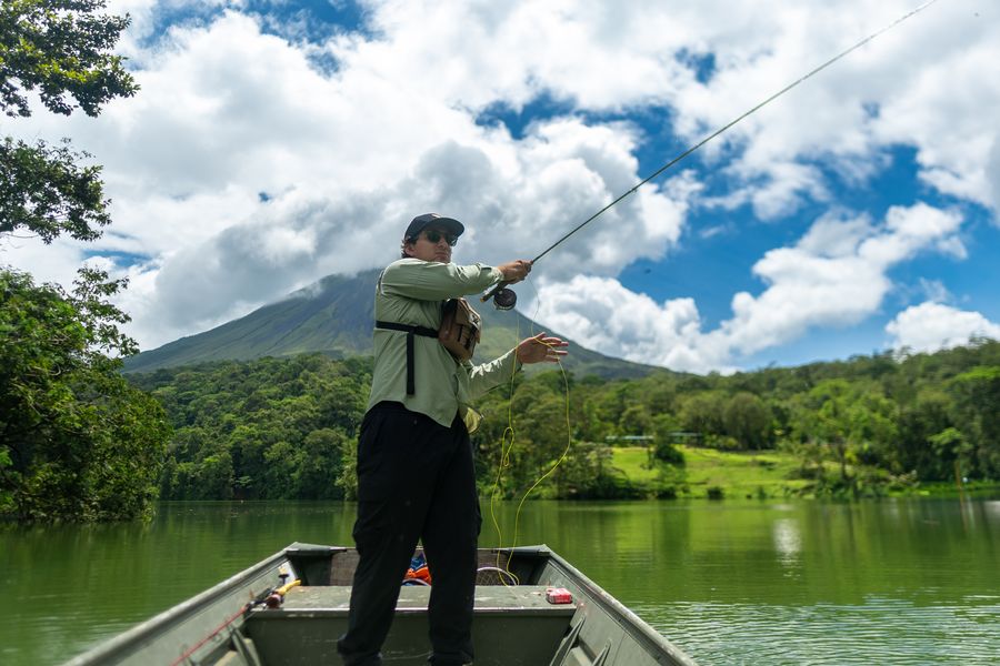 Pescador con mosca lanzando desde una lancha en la Laguna Tabacón, con el Volcán Arenal de fondo